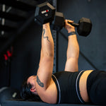 Clad in athletic gear, a person is reclining on a bench and hoisting the SMAI Rubber Hex Dumbbells - (Pair) Apex above their chest. A snake tattoo adorns their arm. The environment resembles a gym with dark walls and various exercise equipment, creating an ideal atmosphere for an intense fitness session.