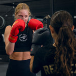 Two women square off in a gym, both in boxing gloves. One wears a black crop top and red/gold SMAI Gold Series Boxing Glove; the other sports black gloves and a shirt with "RESPECT" on the back.
