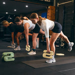 Three women work out in a gym, focusing on single-leg deadlifts using SBELL Chalk 4kg by Sbell Training to enhance grip and performance, all while maintaining balance and proper form on their mats.