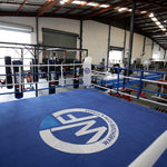 A spacious gym emphasizing fitness and commercial use features an SMAI 6m Competition Boxing Ring in vibrant blue, surrounded by punching bags and exercise equipment beneath a high metal roof. Large windows flood the area with natural light.