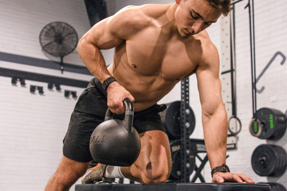A shirtless man in black shorts performs a kettlebell row on a gym bench. Fitness equipment, such as weights and racks, are visible in the background.