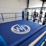 The 6m Competition Boxing Ring from SMAI, with its blue ropes and padding, is the gym's centerpiece. Its free-standing design aligns with the circular "Warehouse Gym & Fitness" logo on the floor. Punching bags are set in the back, while natural light pours in through large windows against white walls.