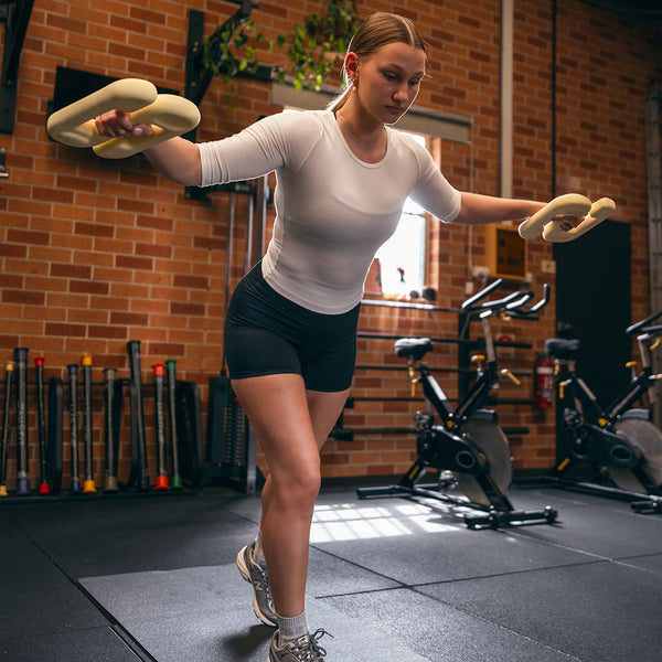 Wearing athletic gear, a woman balances on one leg holding foam weights in a commercial gym with brick walls and Sbell Training’s SBELL Chalk 4kg visible in the background.