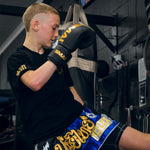 A young athlete in a black t-shirt, blue Muay Thai shorts, and SMAI Gold Series Boxing Glove - Black/Gold trains in the gym, lifting one knee while focusing on their glove for shock protection and improved precision.