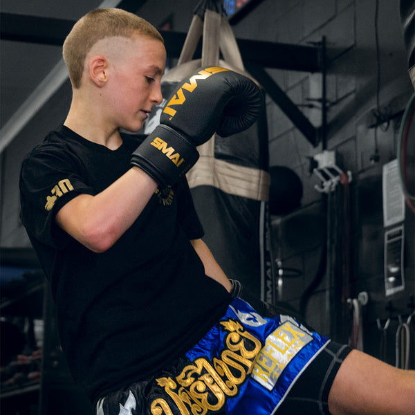 A young athlete in a black t-shirt, blue Muay Thai shorts, and SMAI Gold Series Boxing Glove - Black/Gold trains in the gym, lifting one knee while focusing on their glove for shock protection and improved precision.