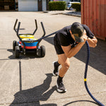 A person in athletic wear pulls the SMAI All Terrain Resistance Sled with ropes across concrete outdoors, showcasing strength training on a sunny day.