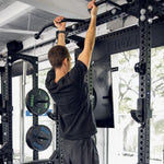 A person in a black workout outfit is doing a pull-up on the SMAI Double Sided Half Rack - Vanta Series in the gym. Weight plates and accessories are visible in the background, with trees framed beautifully through the windows.