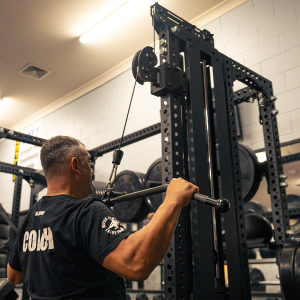 A man in a black shirt uses the SMAI Lat Pull / Low Row Cable Tower Add On 150kg Stack – Vanta Series at the gym, facing away from the camera, with weight plates and a Power Rack system visible in the background.