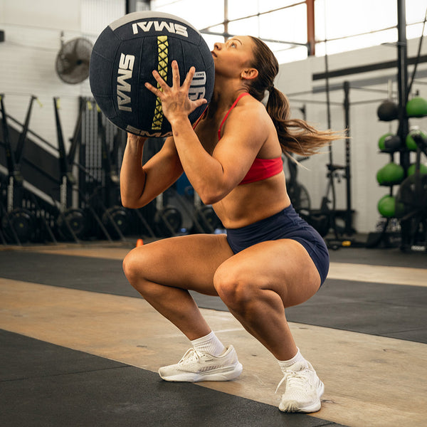 A woman in athletic wear squats in a gym holding an SMAI Wall Balls - Hybrid Range at chest height, with aramid fibre gym equipment and weights in the background, showcasing strength and endurance training.