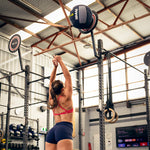 A woman in athletic wear throws a SMAI Wall Balls - Hybrid Range against a wall target in an indoor gym, surrounded by hybrid strength training equipment like rings and weights.