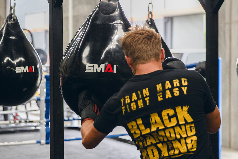 A man wearing a "Train Hard Fight Easy Black Diamond Boxing" shirt punches a black teardrop-shaped punching bag labeled "SMAI" in a gym setting.