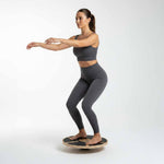 A woman in gray athletic wear demonstrates balance training on the SMAI Balance Board, squatting with arms extended forward against a plain white background.