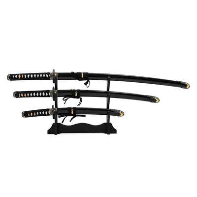 The SMAI Daisho - Carbon 3pc Set with Stand - Shi no Tora features three traditional Japanese swords with black and gold handles, displayed horizontally on a black wooden stand against a white background.