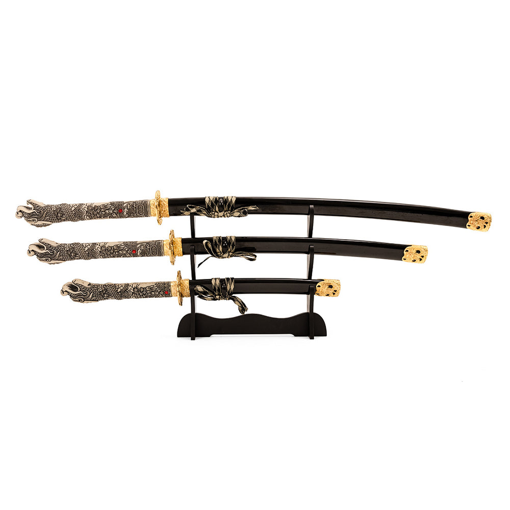 The SMAI Daisho - Highlander Display showcases three decorative Japanese swords with ornate handles and gold accents, arranged horizontally from largest to smallest on a sleek black stand.