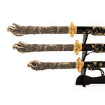 The Daisho - Highlander Display by SMAI showcases three ornate Japanese swords—katana, wakazashi, and tanto—with dragon-detailed handles with red accents and gold guards, all elegantly displayed on a black stand.