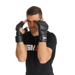 A man wearing black Elite85 MMA Gloves by SMAI, crafted from durable cowhide leather, and a black “SMAI” t-shirt holds his gloved hands near his temples in a fighting stance against a white background.