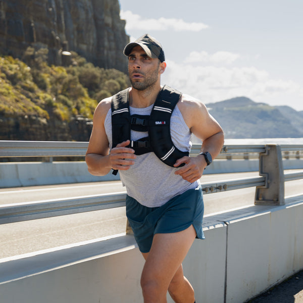 A man in an SMAI Weighted Vest - Olive Green, cap, tank top, and shorts jogs along a seaside bridge with cliffs and hills behind him under a partly cloudy sky—a perfect setting for rucking enthusiasts.