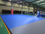 Large indoor industrial space with SMAI Dollamur Flexi Connect Gym Flooring Roll in Royal Blue, featuring advanced athletic foam, surrounded by machinery and workstations under high ceilings with exposed beams and fluorescent lighting.