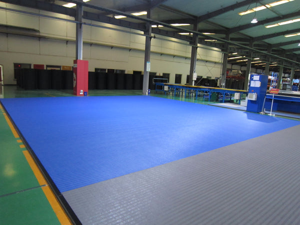 Large indoor industrial space with SMAI Dollamur Flexi Connect Gym Flooring Roll in Royal Blue, featuring advanced athletic foam, surrounded by machinery and workstations under high ceilings with exposed beams and fluorescent lighting.
