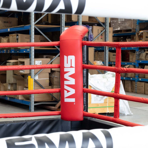 A warehouse-like space houses a corner of a boxing ring adorned with red SMAI-branded padding, surrounded by shelves stacked with boxes. This setup includes SMAI Boxing Ring Rope Spacers for optimal rope stability, featuring an easy-install design.
