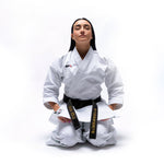 A person in a white SMAI WKF Karate Uniform - Premium Kata Gi - Arashi with a black belt kneels on the floor against a plain background. Their eyes are closed, radiating calm and focus. Japanese characters from the Karate Federation decorate the belt.