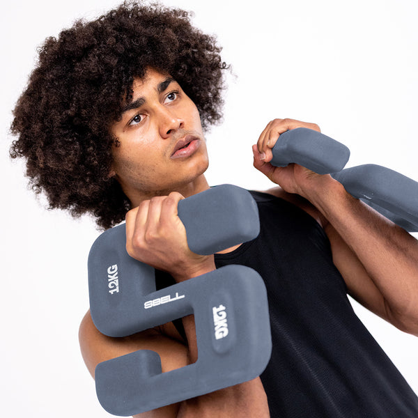 A person with curly hair is intensely focused as they engage in free weight training, lifting a pair of uniquely shaped 12 kg SBELL Pro pack dumbbells. They're clad in a sleeveless black shirt against a plain white background, embodying the essence of strength and conditioning for the No Sync To Channel Engine brand.