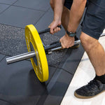 Wearing black shorts and sneakers, a person grips the SMAI The Yard - Core Trainer / Landmine Parallel Row Handle with a large yellow weight plate, preparing to lift on the gym floor.