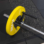 A silver barbell with a yellow 15 kg plate rests on a speckled gym mat, featuring the SMAI The Yard - Core Trainer / Landmine Parallel Row Handle for enhanced grip.