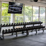 A gym features the SMAI Commercial Dumbbell Set with Rack 5-60kg, neatly arranged in front of large windows. Above the setup, a screen displays "Performance Everyday." Visible outside are trees and a building, while rubber mats cover the floor. The rubber-coated weights contribute to the professional atmosphere.