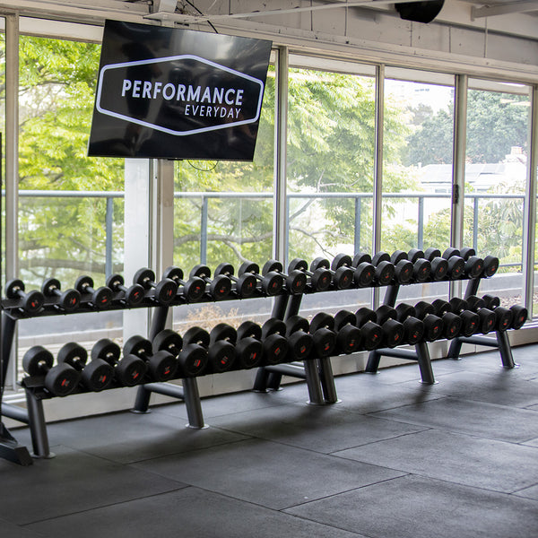 A gym features the SMAI Commercial Dumbbell Set with Rack 5-60kg, neatly arranged in front of large windows. Above the setup, a screen displays "Performance Everyday." Visible outside are trees and a building, while rubber mats cover the floor. The rubber-coated weights contribute to the professional atmosphere.