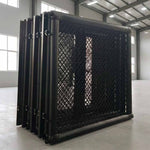 The SMAI MMA Cage - 5m Fold Away, a large black metal mesh structure, stands folded inside a spacious industrial warehouse with high ceilings and large windows.
