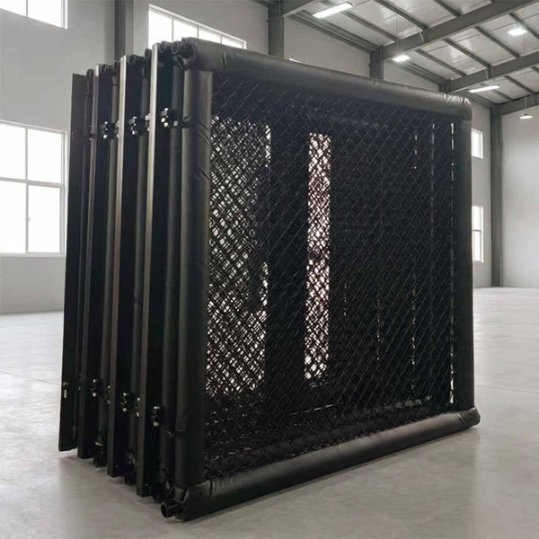 The SMAI MMA Cage - 5m Fold Away, a large black metal mesh structure, stands folded inside a spacious industrial warehouse with high ceilings and large windows.