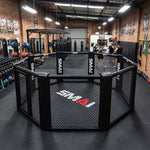 A gym with brick walls features an SMAI MMA Cage - 5m Fold Away in the center, surrounded by weights, punching bags, and equipment. Several people are blurred in the background near the cage.