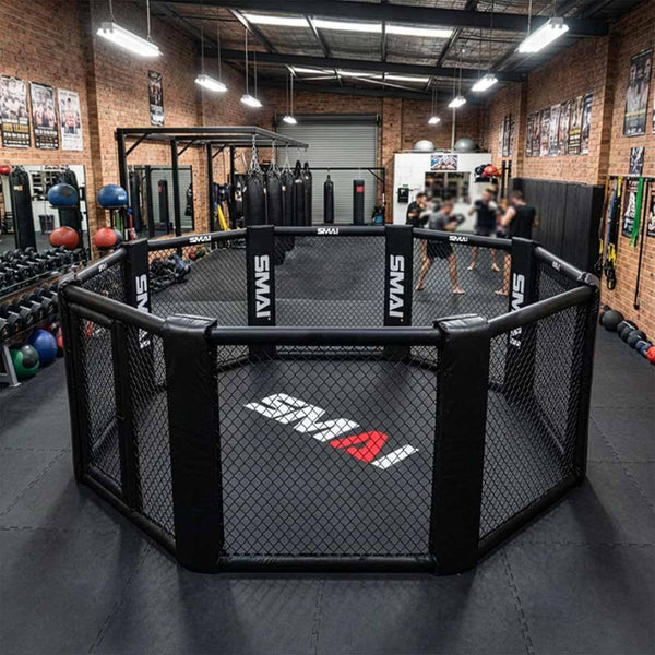A gym with brick walls features an SMAI MMA Cage - 5m Fold Away in the center, surrounded by weights, punching bags, and equipment. Several people are blurred in the background near the cage.
