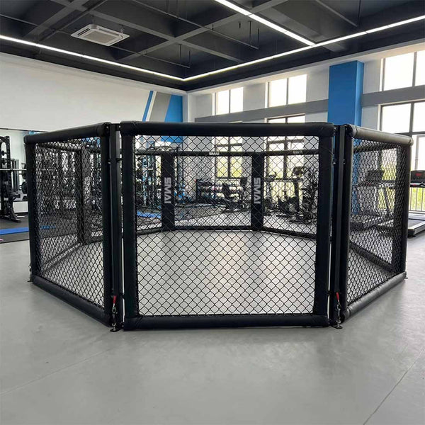 A black SMAI MMA Cage - 5m Fold Away with chain-link fencing stands in a bright gym, featuring large windows and exercise equipment in the background.