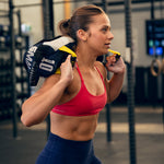 A woman wearing athletic wear lifts the SMAI Sand Bags - Hybrid Range onto her shoulders in a gym, looking focused and determined. Exercise equipment and gym racks appear in the background, emphasizing strength and endurance training.
