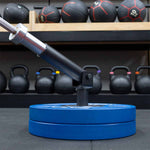 A SMAI The Yard - Drop In Landmine / Core Trainer holds a barbell anchored by two blue plates, with kettlebells and medicine balls displayed on shelves in the background.