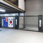 A martial arts training area features the SMAI MMA Cage Wall - Single Panel, a black chain-link fence that resembles a reinforced cage mesh. Displayed prominently are the Australian and South Korean flags, while in the background, a partially closed garage door complements the ceiling's overhead lighting.