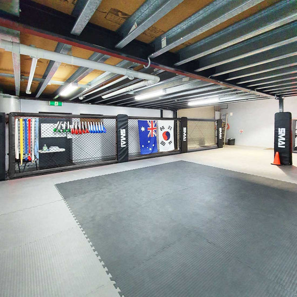 A martial arts gym features a mat area and punching bags, with an array of colored belts showcased on a rack. The space is accented by modular cage walls, including the SMAI MMA Cage Wall - Single Panel, adorned with flags from Australia and South Korea. The exposed pipes on the ceiling contribute to the gym's industrial aesthetic.