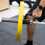 A person stands on gym flooring, wearing a smartwatch, preparing to exercise with the SMAI "The Yard - Single Barbell Handle" landmine accessory and a barbell with a yellow weight plate in a gym.