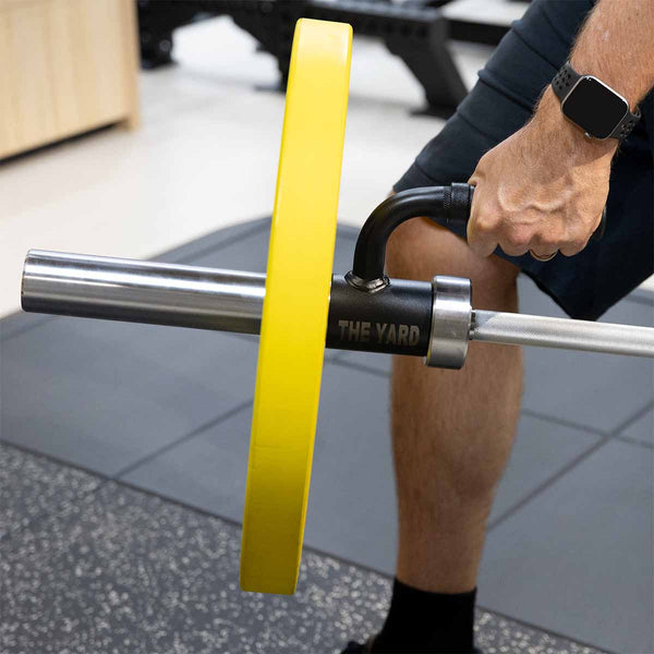 A person stands on gym flooring, wearing a smartwatch, preparing to exercise with the SMAI "The Yard - Single Barbell Handle" landmine accessory and a barbell with a yellow weight plate in a gym.