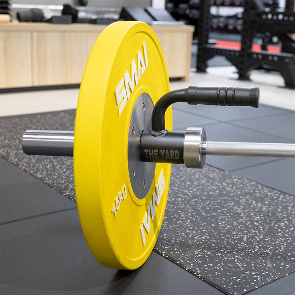 A yellow 15 kg SMAI weight plate and a black "The Yard" clamp secure a barbell resting on rubber mats with the SMAI Single Barbell Handle - Landmine Accessory nearby, gym equipment in the background.