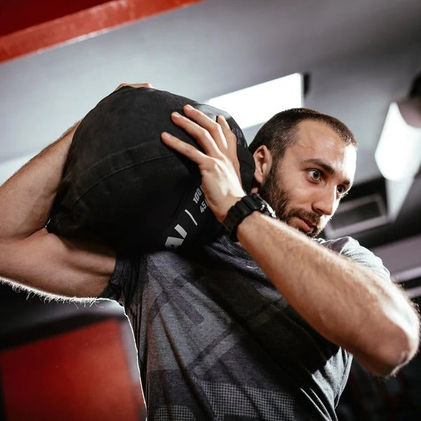 A man with a beard lifts the SMAI Strongman Sandbag onto his shoulder during an intense gym workout, focusing on proper form.