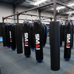 A gym showcases an array of hanging punching bags in a spacious, well-lit area with blue flooring. Each bag is supported by a sturdy 3-meter Mammoth Rig Cross Beam made of high-gauge steel tubing, arranged in rows and ready for training sessions. The ceiling features exposed beams and metal fixtures.