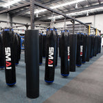 A spacious gym showcases multiple black punching bags branded "SMAI," suspended from a Mammoth Rig Cross Beam - 3m. The facility is accented with a blue-speckled rubberized floor and an industrial-style ceiling, with various other fitness equipment seen in the background.