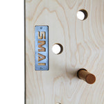 Close-up of the SMAI Monster Peg Board: light marine ply with round holes, a metal "SMAI" plate, and a wooden peg inserted—perfect for load-bearing wall mount setups and intense peg board workouts.