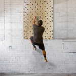 Wearing athletic clothing, a person climbs the SMAI Monster Peg Board, made of marine ply and fixed to a white brick wall, gripping pegs with their hands and supporting their weight with their legs.